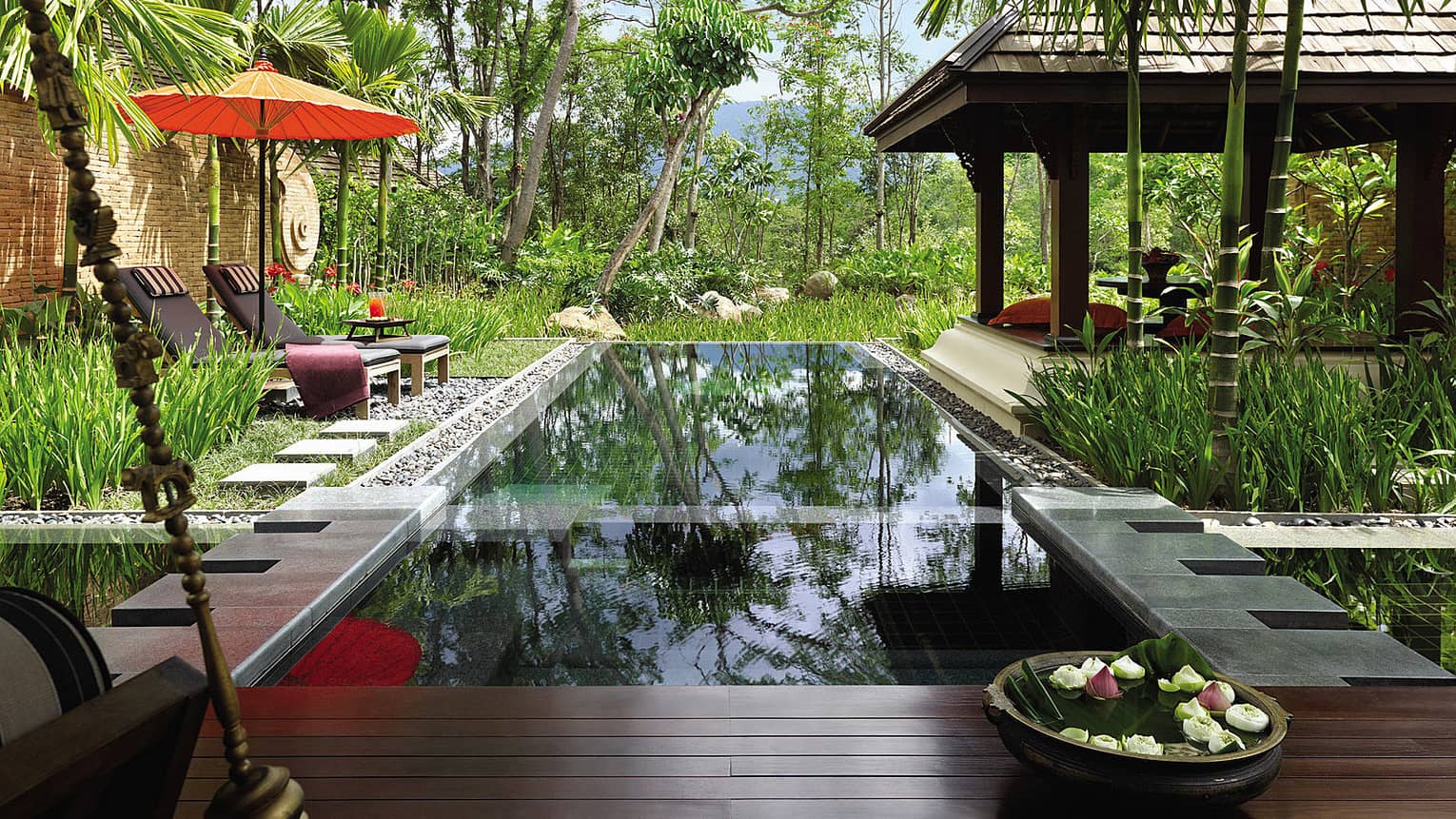 Bowl with fresh flowers by private swimming pool on villa patio with lounge chairs orange under umbrella