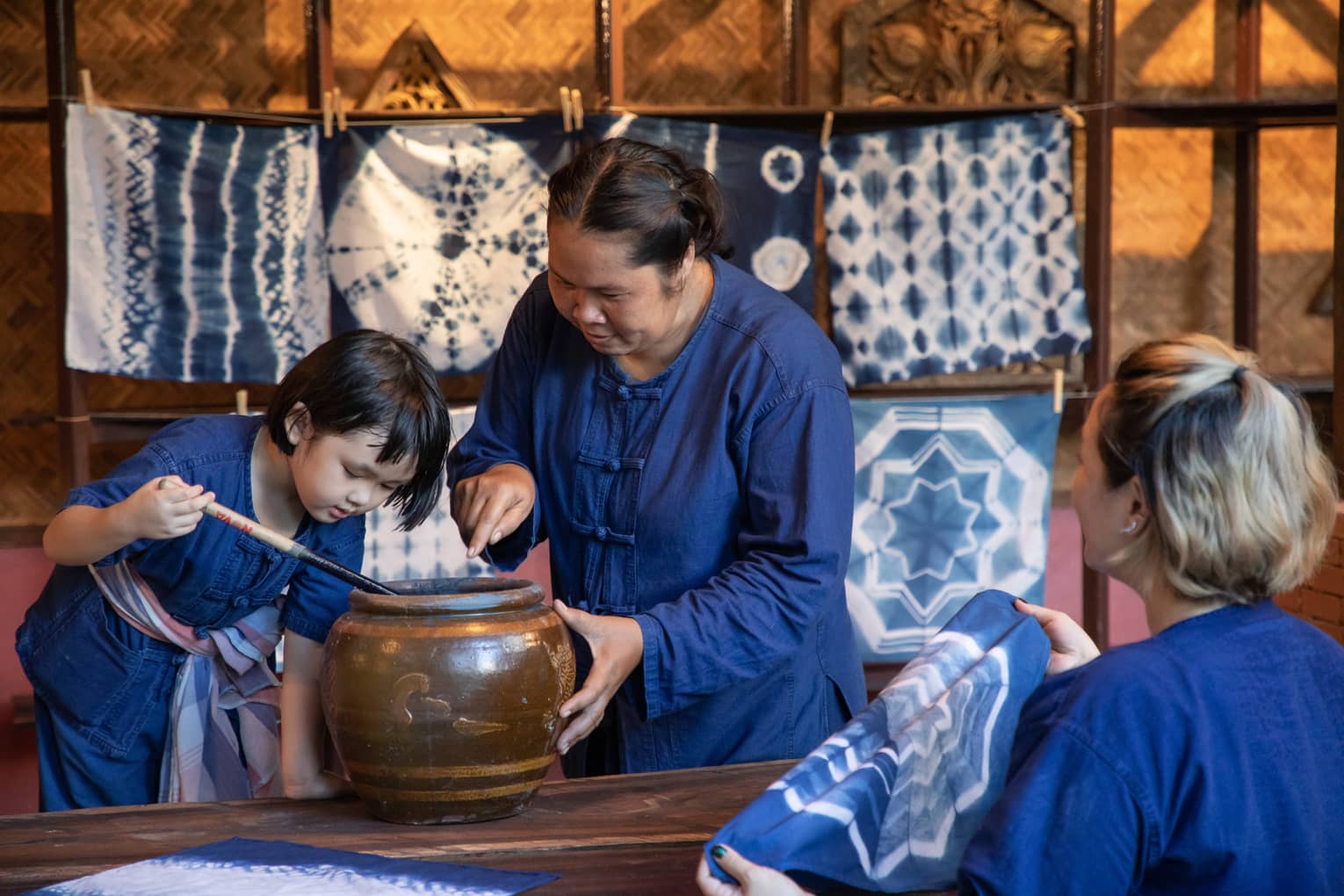 A woman teaching a child how to dye fabric using indigo, with tie-dyed cloths hanging in the background in a traditional workshop.