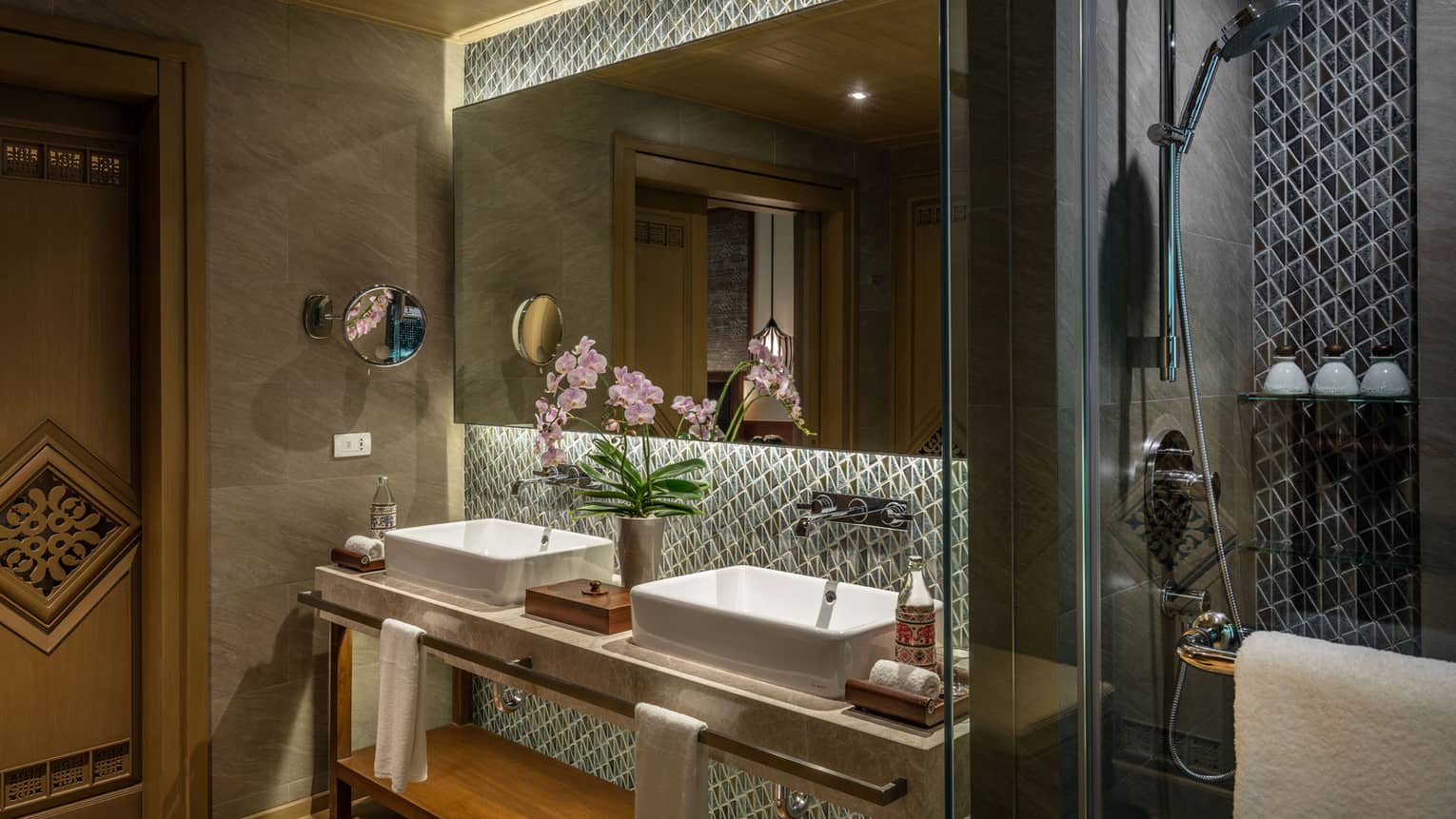 Luxurious bathroom with double vanity, orchid decor, large mirror, geometric tile backsplash, glass-enclosed shower and warm lighting