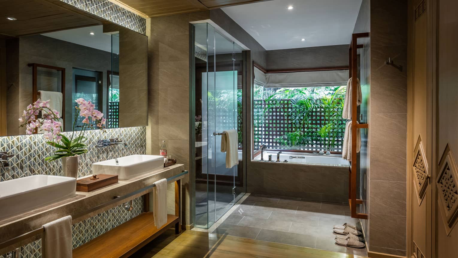 Luxurious bathroom with double sinks, orchid decor, glass shower, bathtub with garden view, geometric tile backsplash and soft lighting