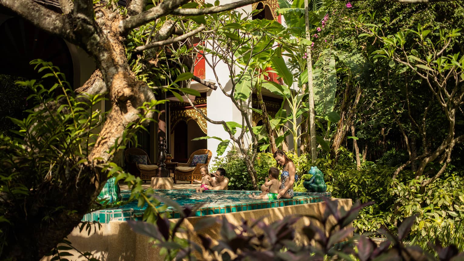 Couple with two babies sit on edge of private pool in front of bungalow by garden