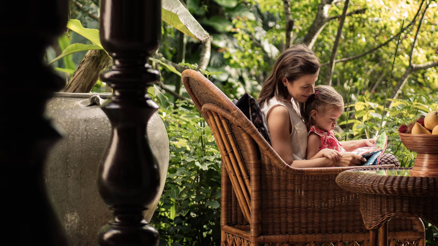 Woman read to young girl on her lap in large wicker armchair on patio by trees