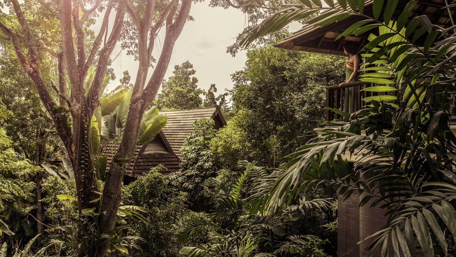 Woman on villa balcony looks out over tropical palms, trees, roof of next building 