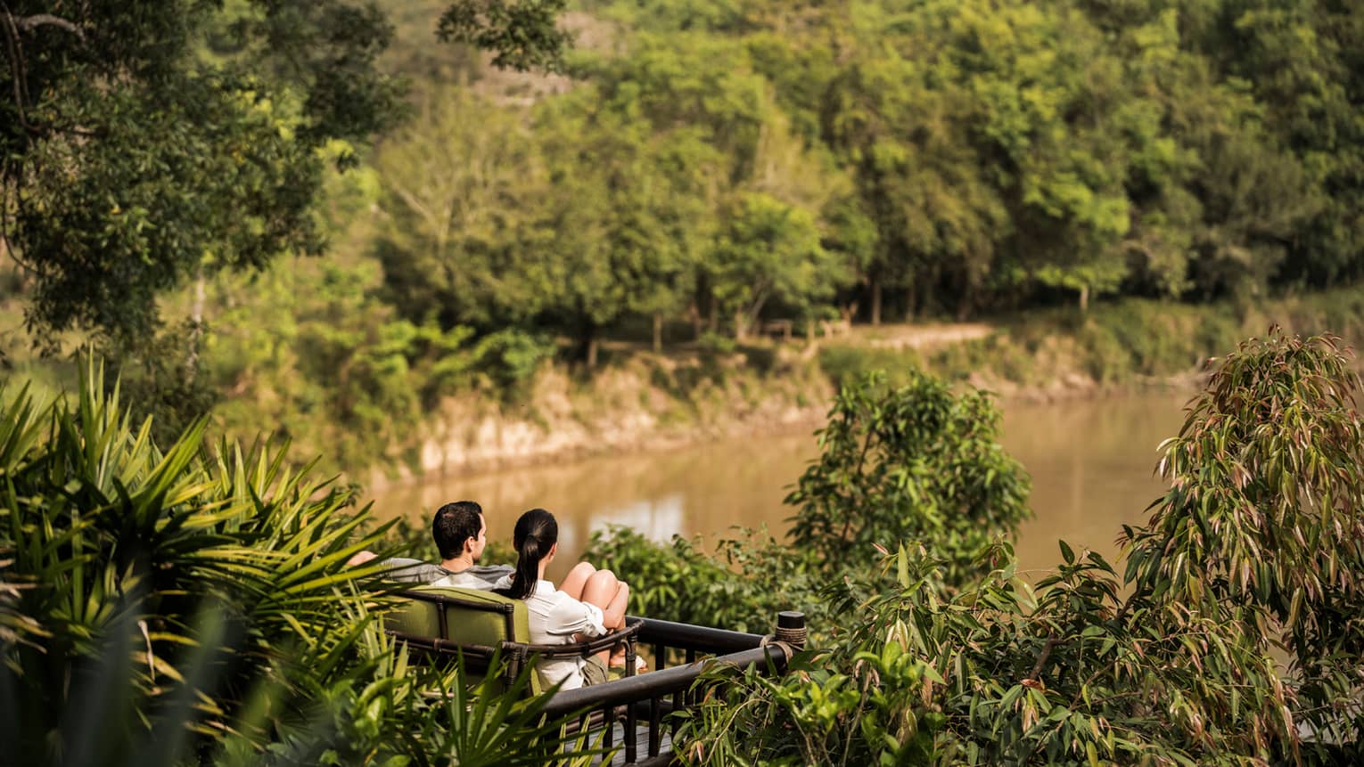 Couple sits on wood bench surrounded by tropical plants, overlooking river
