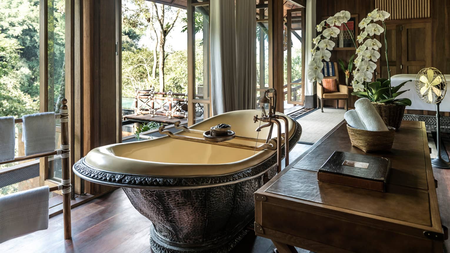 Free-standing copper bathtub in front of windows, wood dresser with rolled white towels