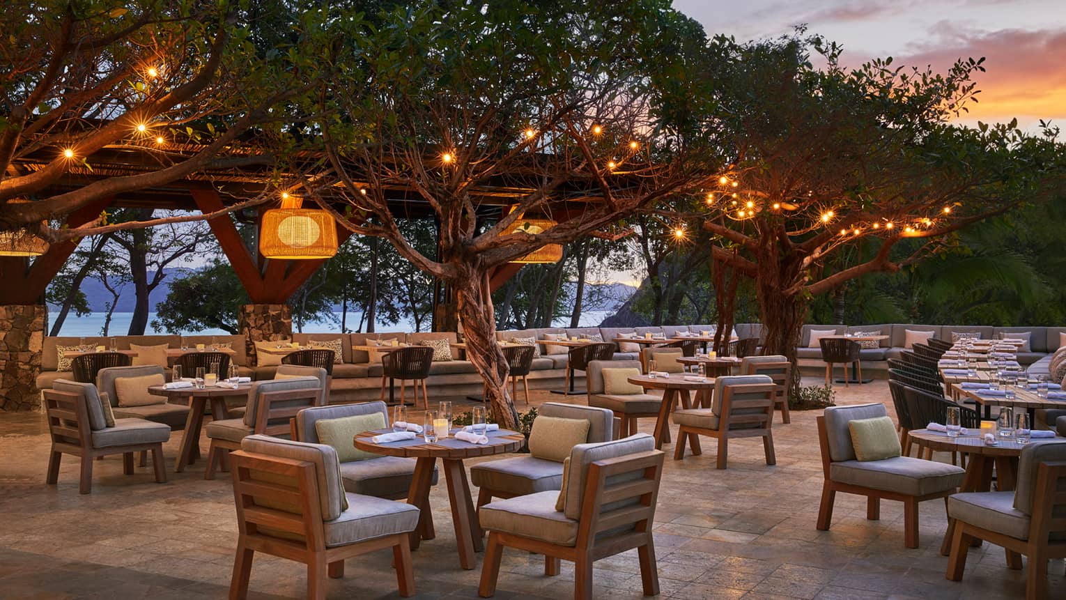 Patio tables, chairs with plush cushions on patio under trees with hanging lights, at sunset
