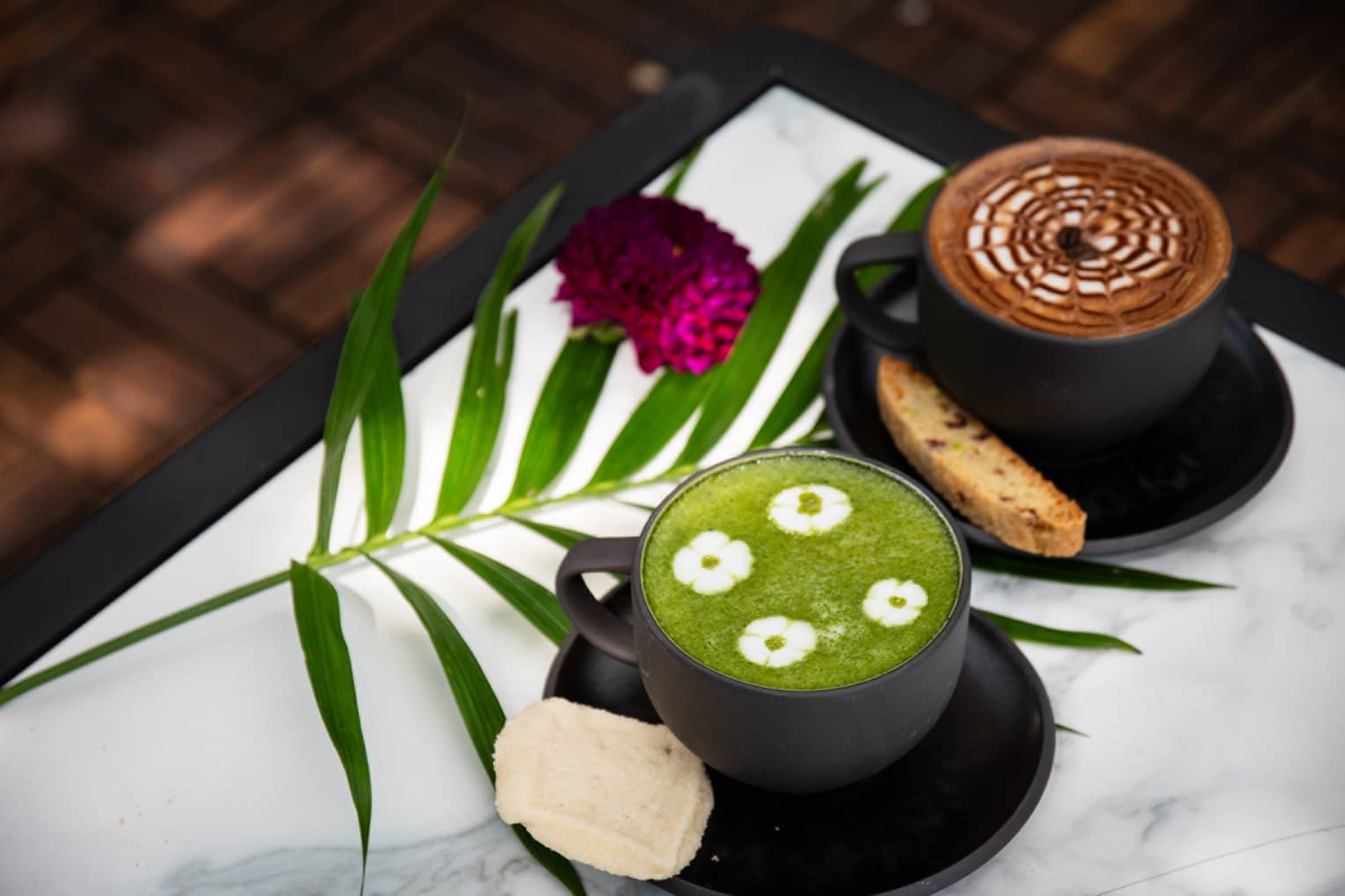 A black mug of coffee with a latte and brown-and-white foam and another black mug with tea with green foam and four white floral patterns