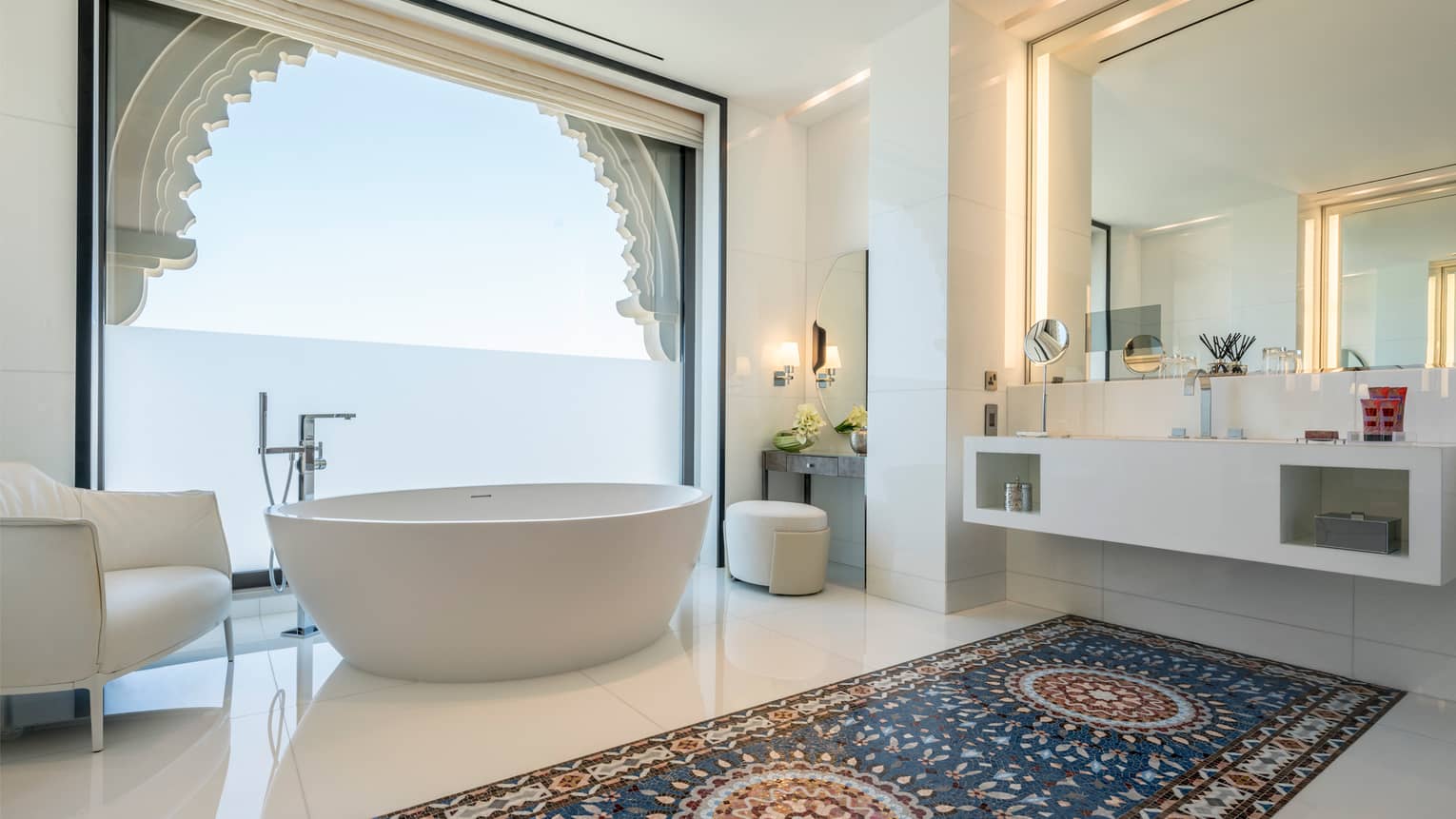 Bathroom with white spa tub and armchair in front of tall window and vanity, white floors with Arabic-style tiles