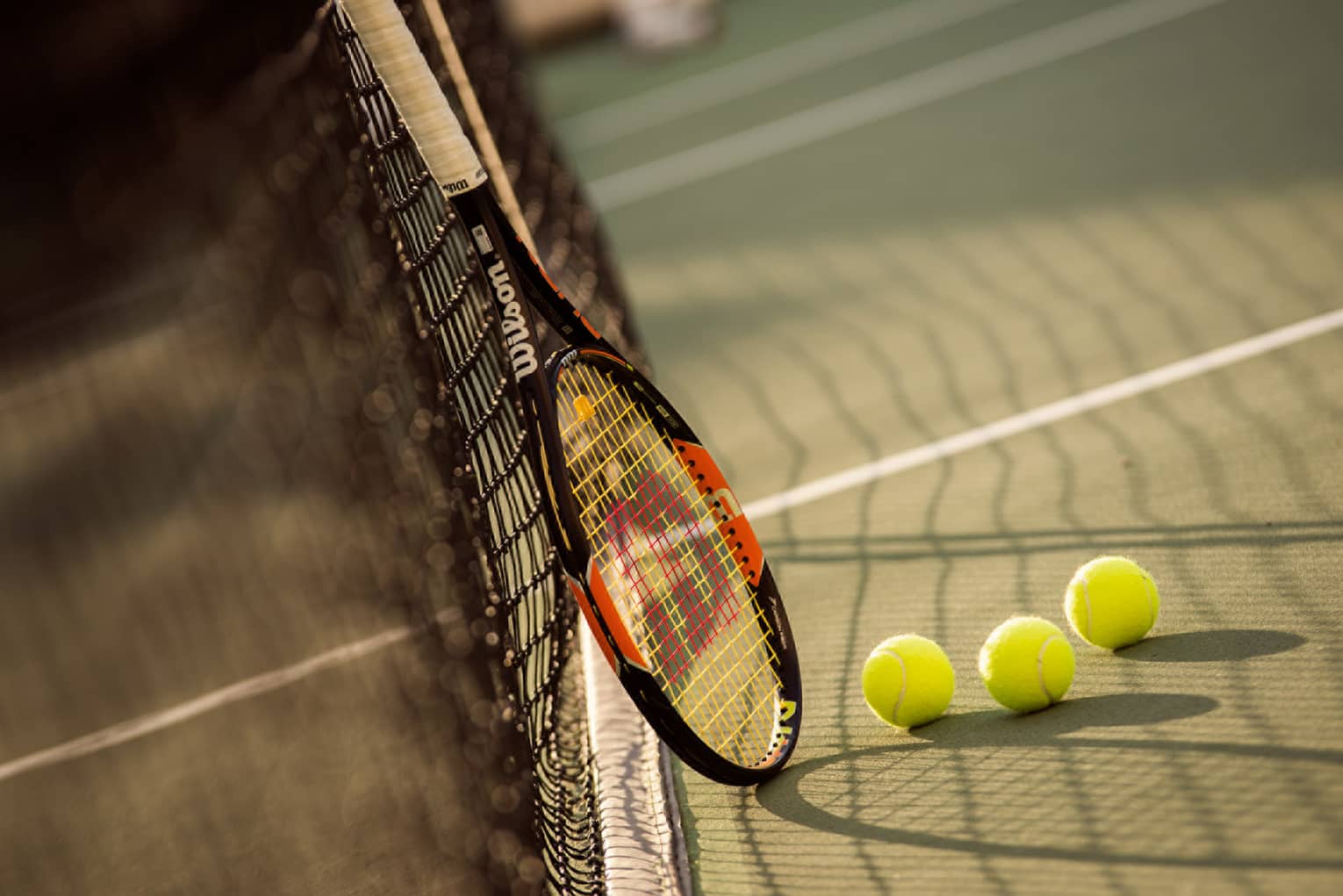 Red and black tennis racket leans against net by green tennis balls on court