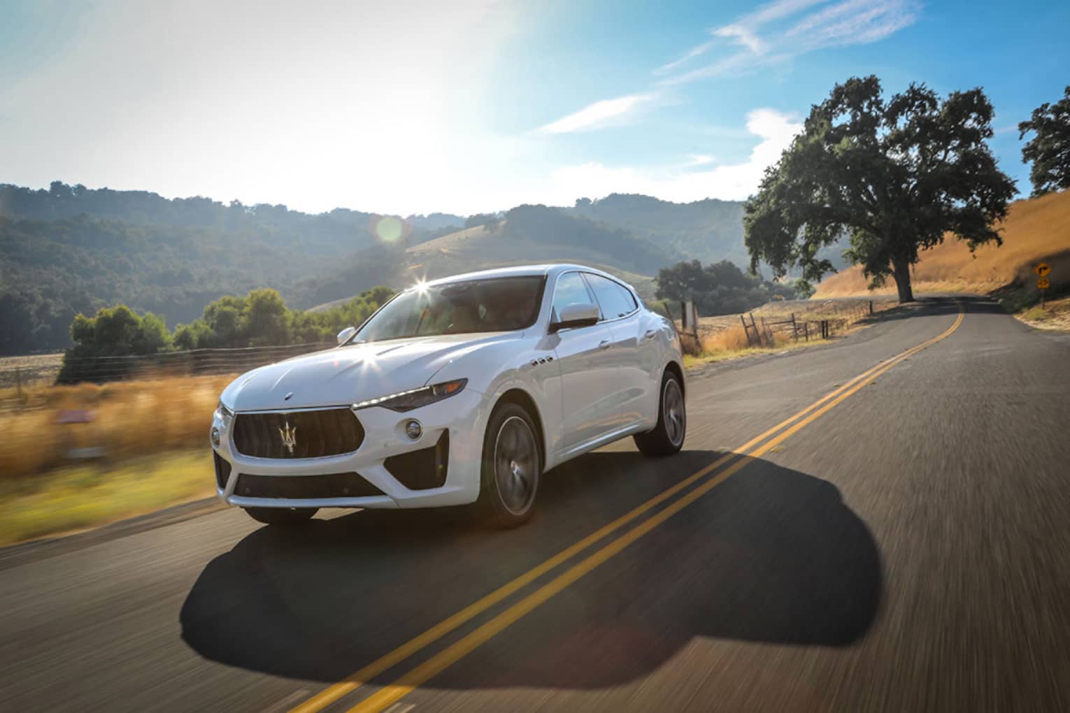 White Maserati Levante SUV on two-lane roadway on sunny day, with blue skies, green hills, fields and trees in background