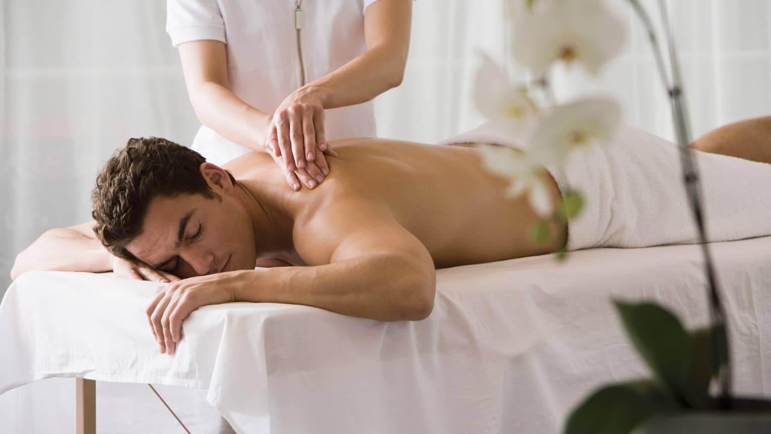Man lays on white table with eyes closed as masseuse massages his bare shoulders