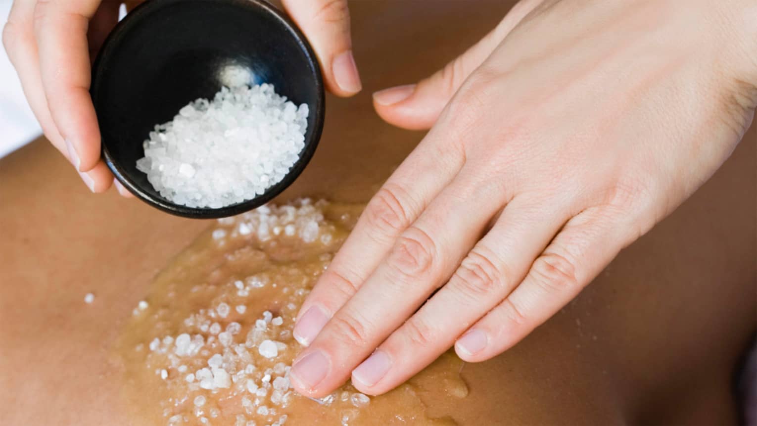 Close-up of large spa salts in bowl, hand rubbing crystals on bare back