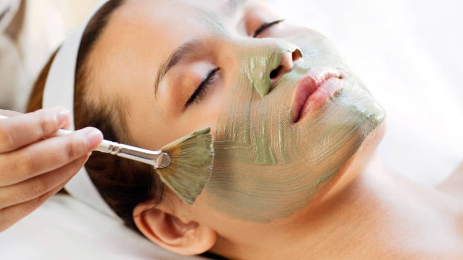 Spa staff holding brush paints green mask on woman's cheeks, chin, nose