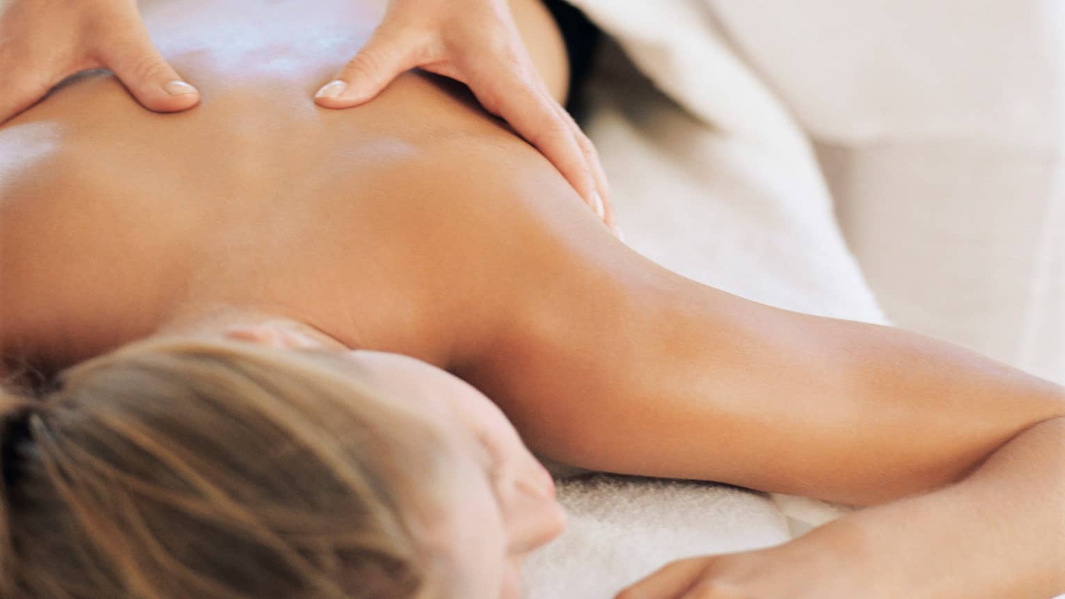 Woman lays face-down on massage table under white blanket as hands massage her bare shoulders
