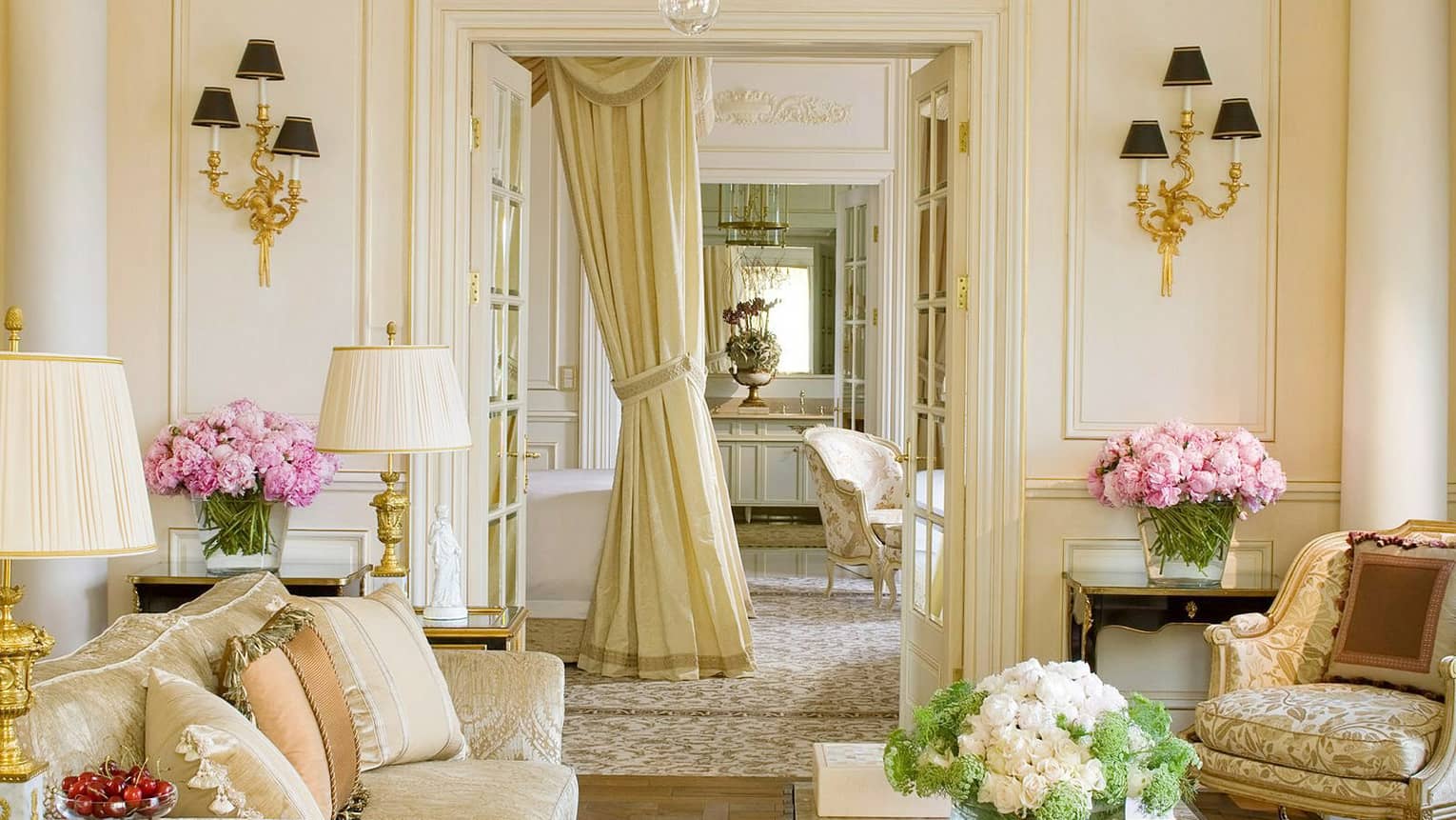 Royal suite living space with cream-coloured walls, gold accents, floral arrangements, sofa, armchair and luxurious drapery leading into an adjoining room