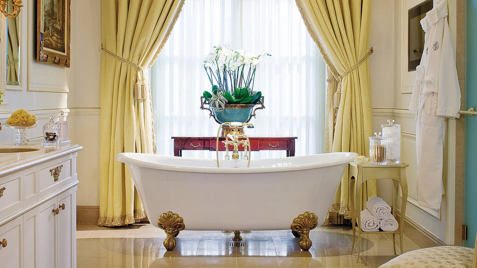 Royal suite bathroom featuring a freestanding clawfoot bathtub with gold fixtures, elegant yellow drapes framing a tall window and white bathrobe hanging nearby