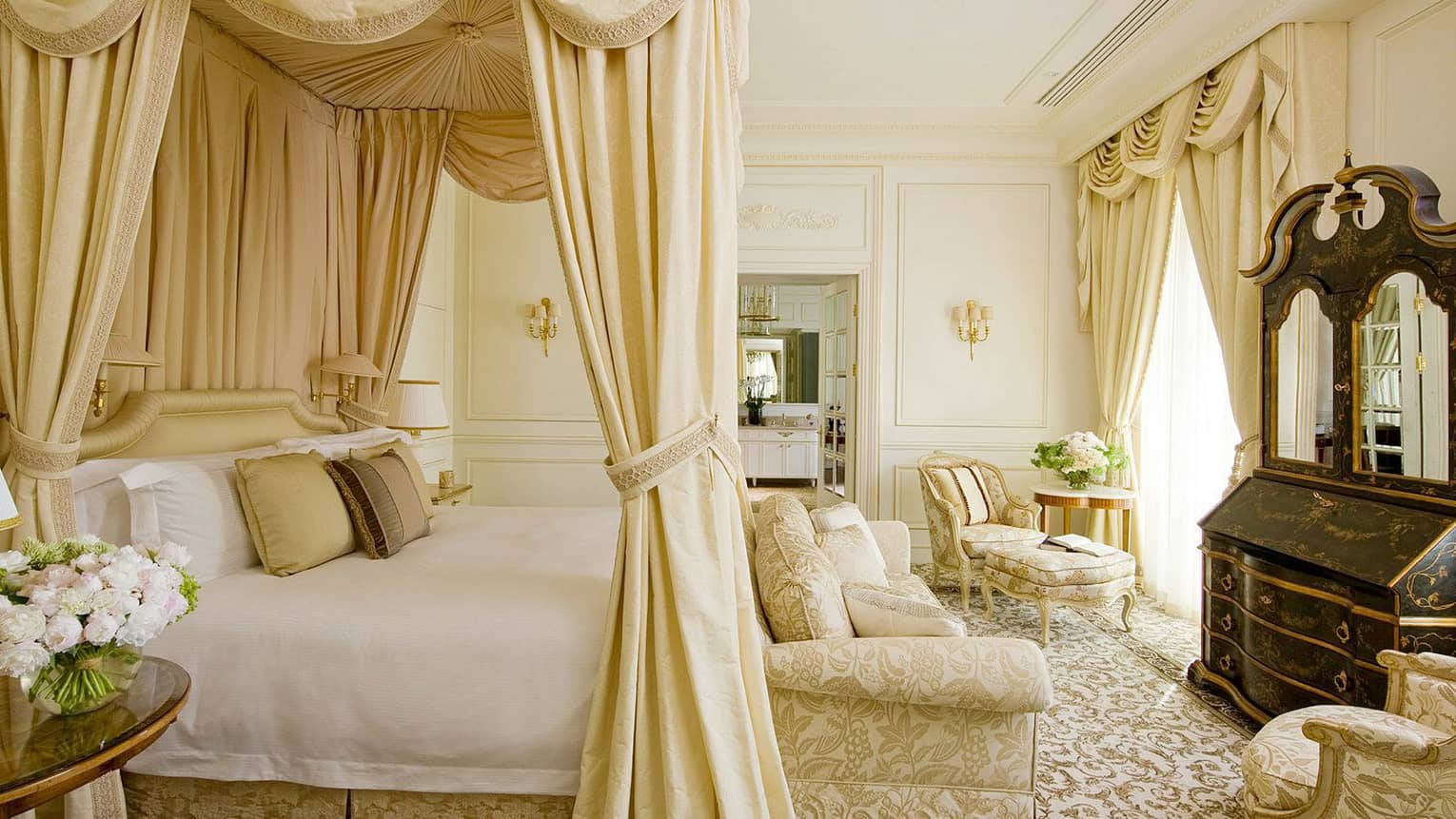 Royal suite bedroom featuring a grand four-poster bed with gold drapery, elegant upholstered chairs, sofa, black lacquered armoire and classic cream décor