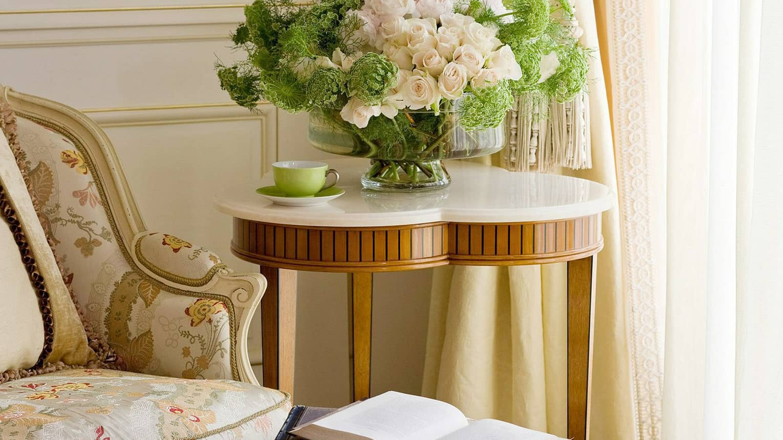 Floral upholstered armchair next to a side table holding a bouquet of white roses and green foliage, with an open book and a green teacup placed nearby