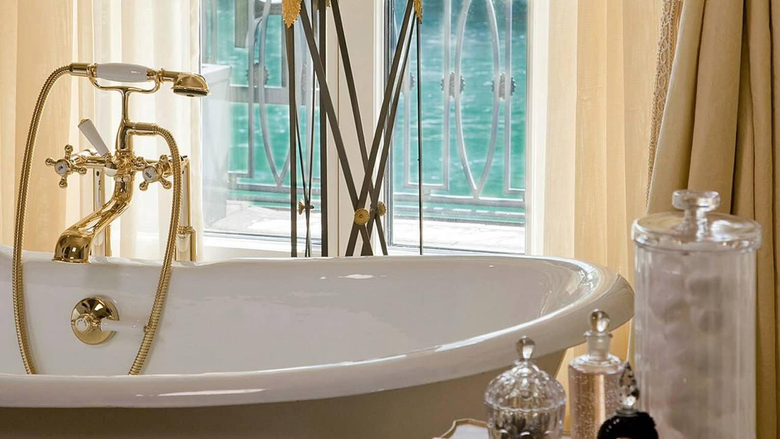 Close-up of a luxurious freestanding bathtub with gold fixtures, framed by sheer curtains and offering a view of the water through a balcony window