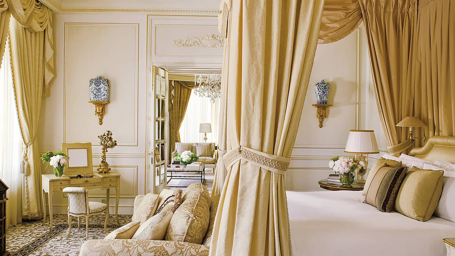 Opulent suite bedroom with a four-poster bed, gold drapes, ornate furnishings, a small dressing table and a sitting area in the adjoining room