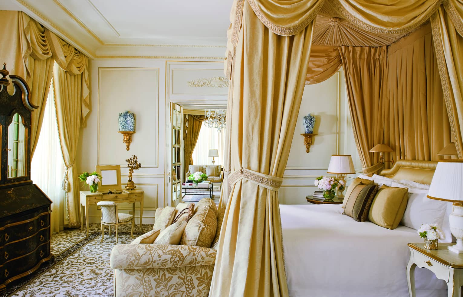 Opulent suite bedroom with a four-poster bed, gold drapes, ornate furnishings, a small dressing table and a sitting area in the adjoining room