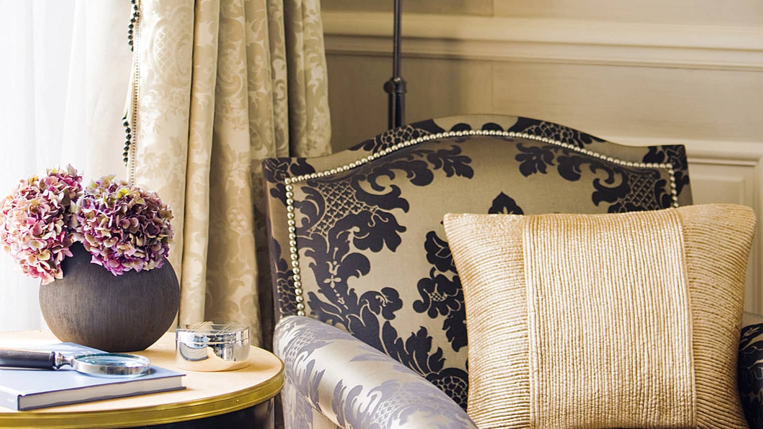 Close-up of an elegant armchair with a bold damask pattern, paired with a gold textured cushion, next to a side table holding a vase of pink flowers