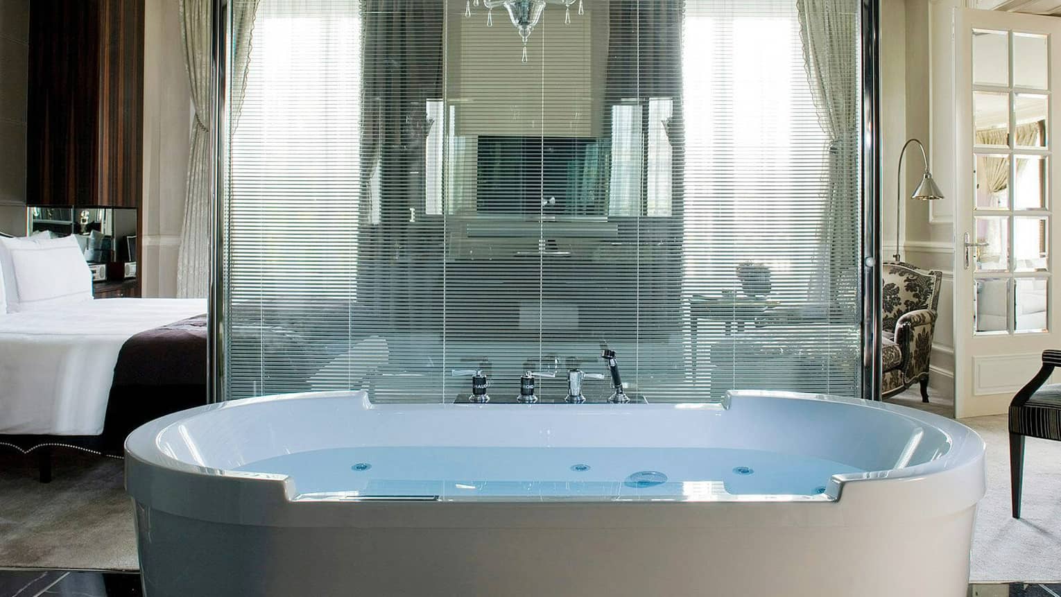 Presidential suite bathroom with a large soaking tub, glass partition with blinds and a view into a luxury bedroom featuring neutral tones and elegant furniture
