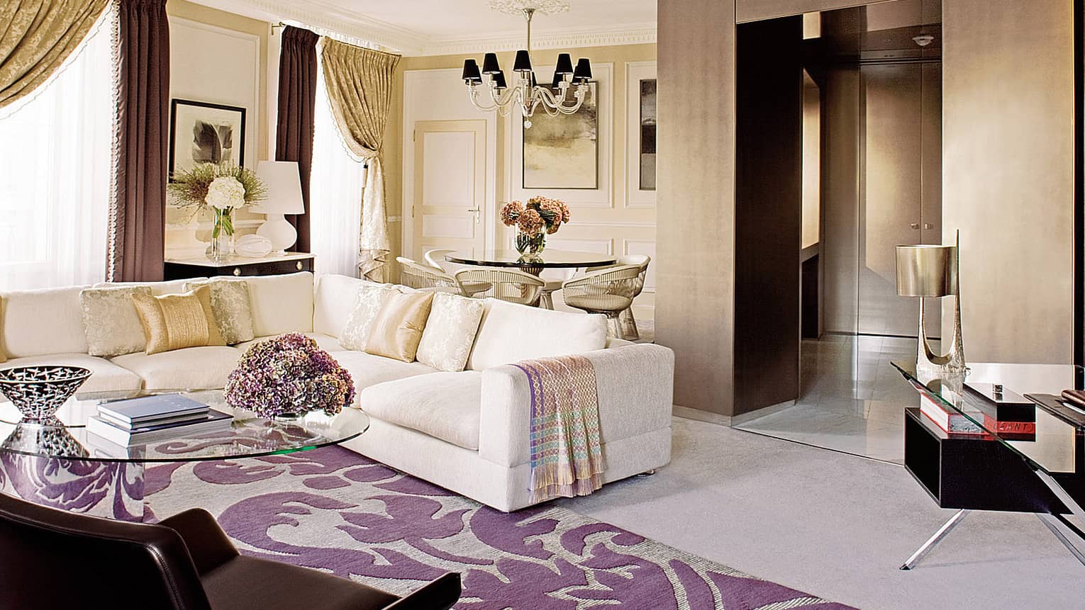 Luxury living room with a cream sectional sofa, purple patterned rug, glass coffee table and a dining area with a glass-top table, flowers and chandelier 