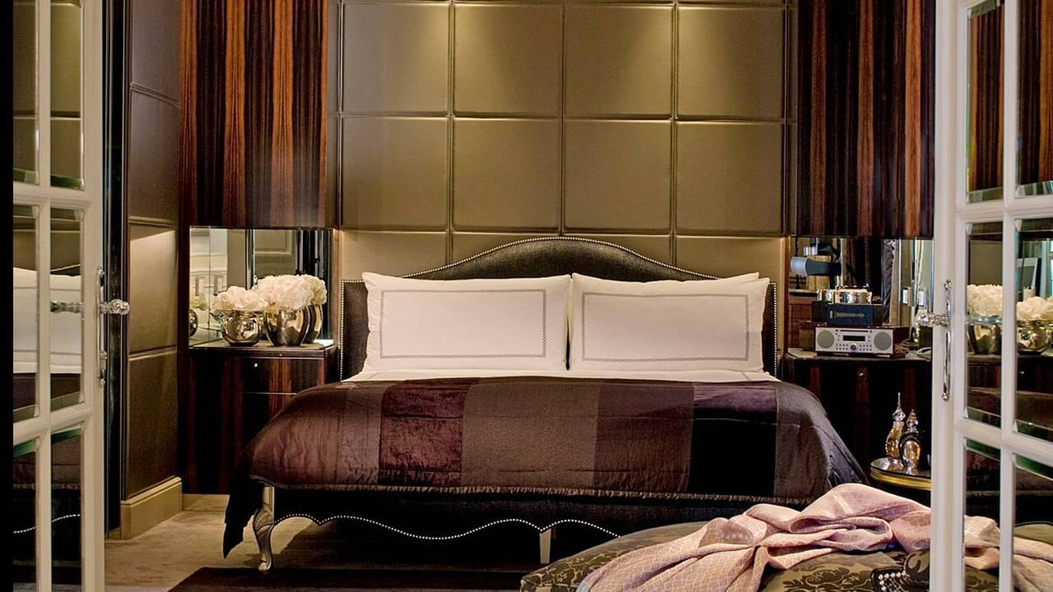 Luxury bedroom with a tufted headboard, dark wood panelling and rich brown bedding, complemented by mirrored side tables and modern décor accents