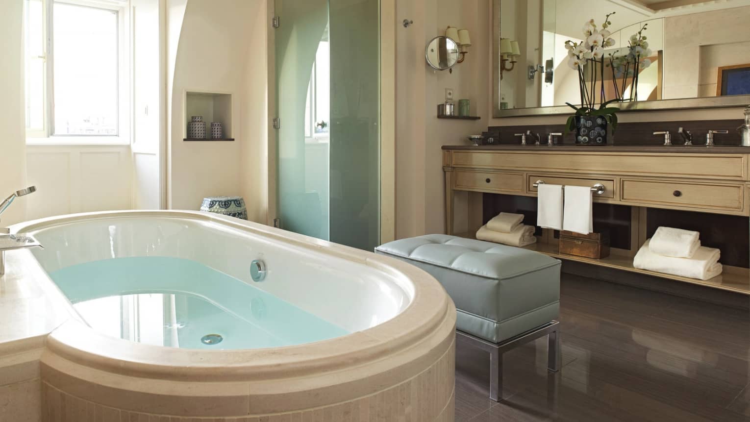 Luxury hotel bathroom with a large oval bathtub, double sink vanity, glass shower, upholstered bench and a window providing natural light