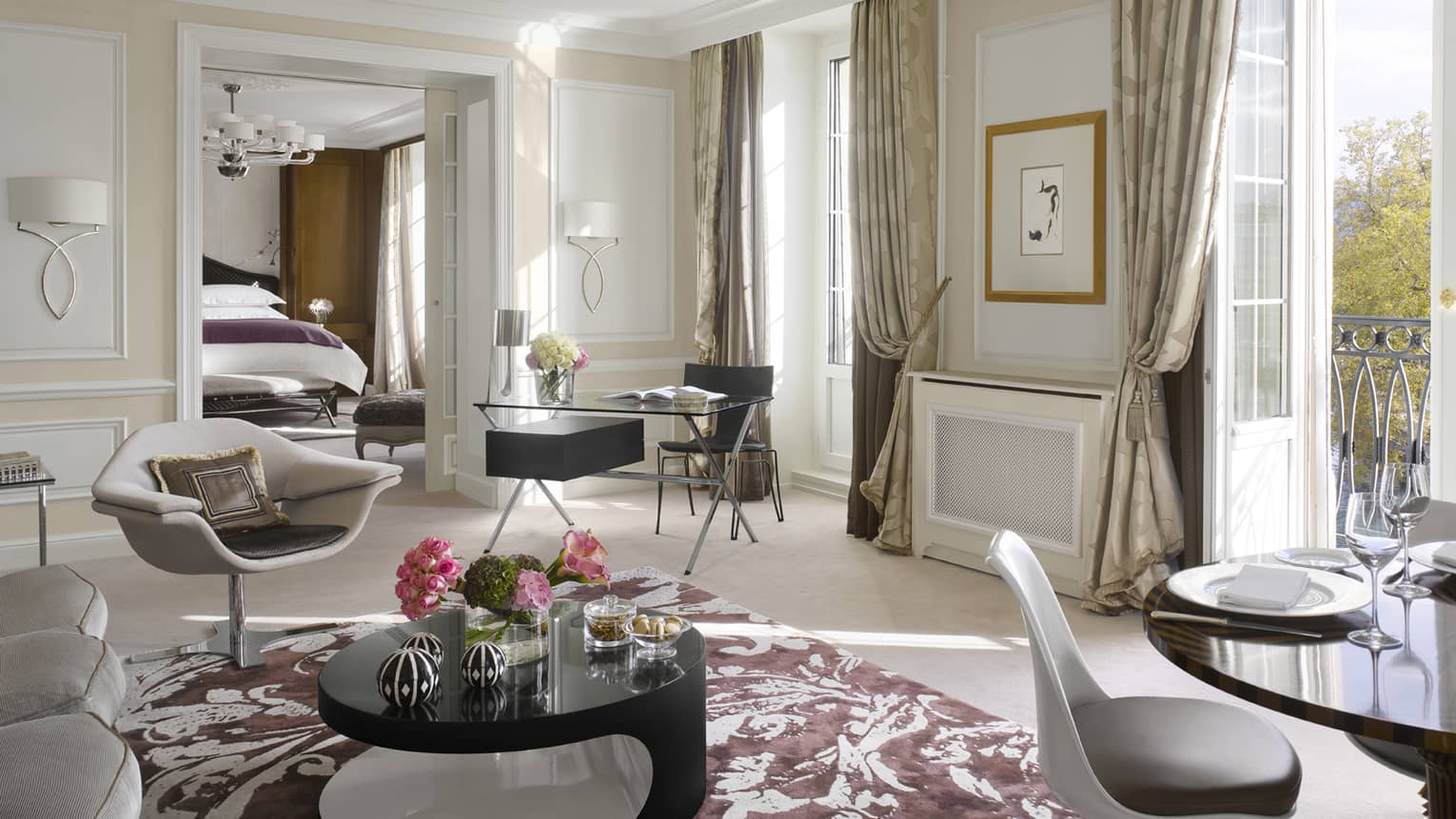 Luxury hotel suite living area with a modern armchair, black desk, round coffee table, dining table and balcony access, leading to a bedroom with a king bed