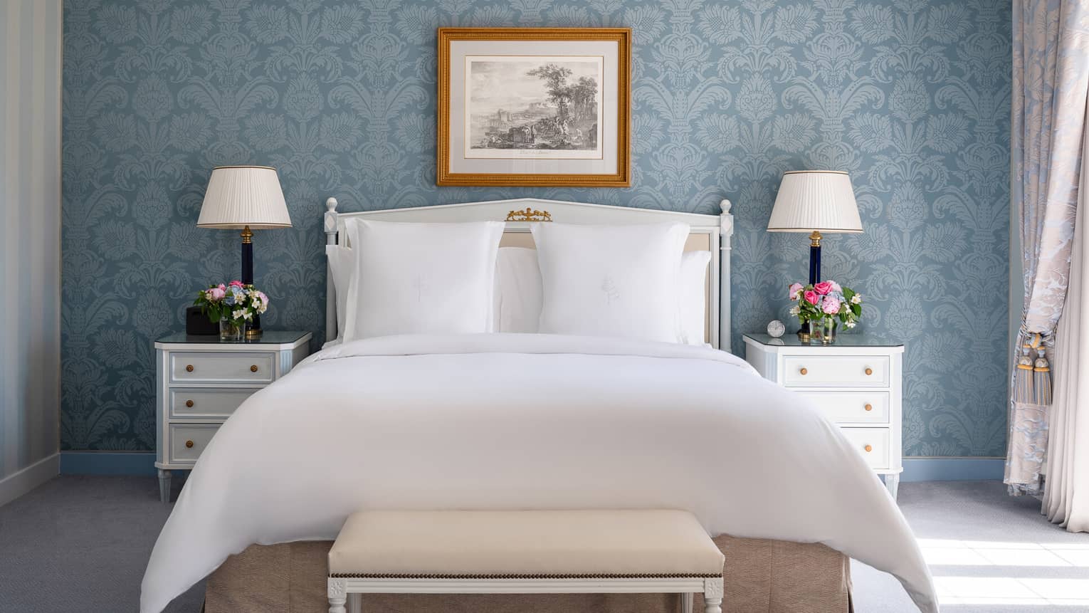 Elegant hotel bedroom with a king bed, white bedside tables with lamps and flowers, bench, blue floral wallpaper and framed artwork 