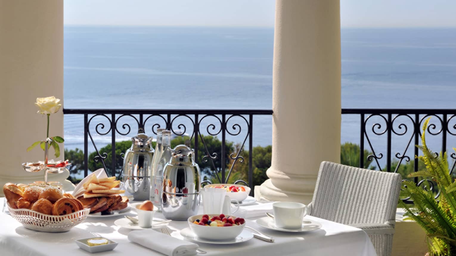 Brunch is arranged on a square table with white table cloth, white whicker chair on balcony overlooking the sea