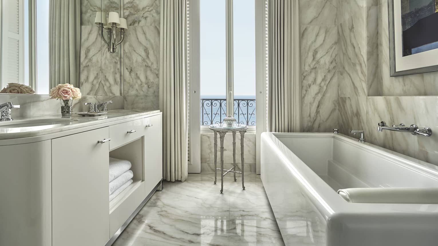 Full-marble hotel bathroom with double vanity, plate mirror, soaking tub and Mediterranean Sea views out tall window flanked by curtains