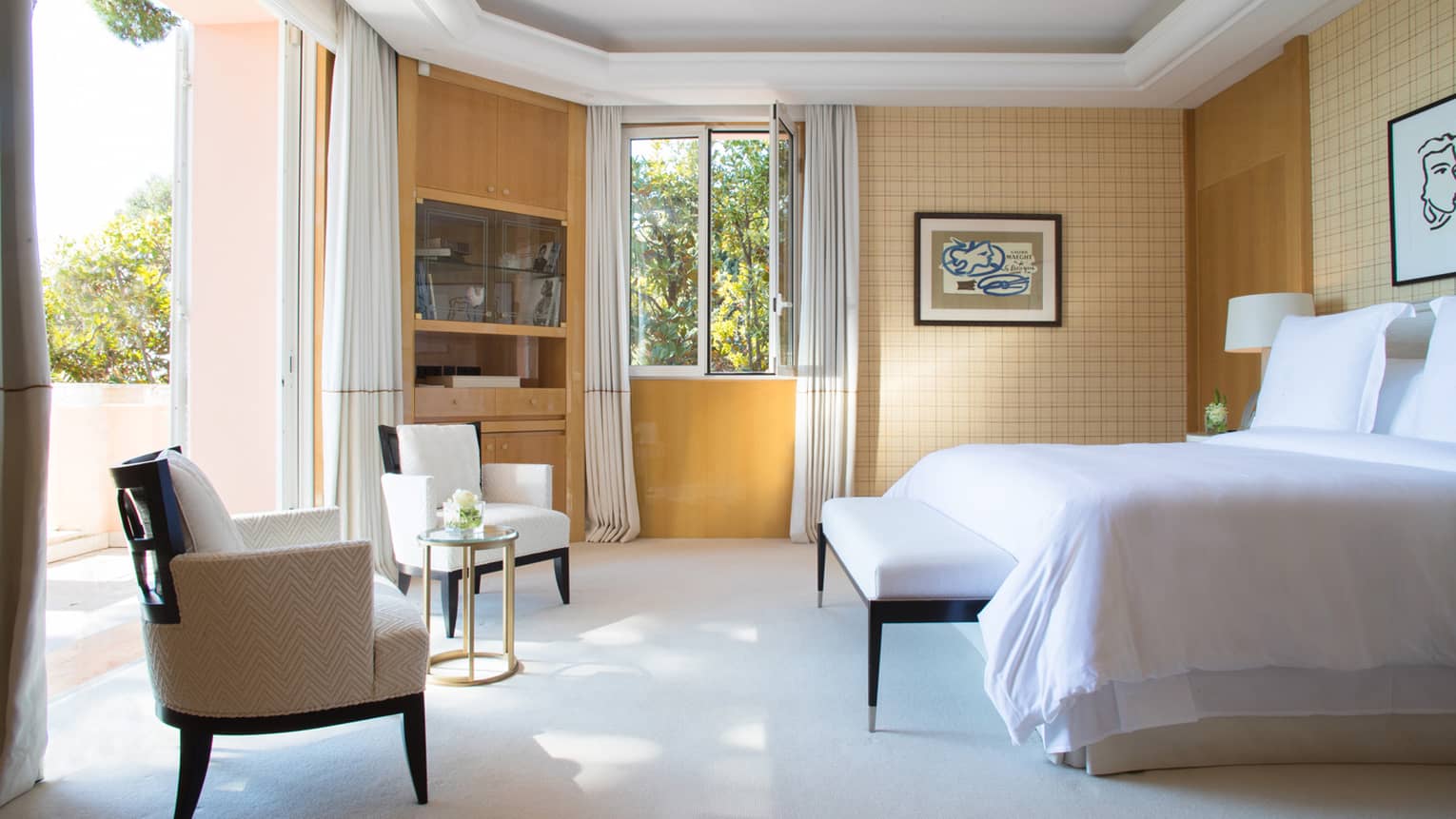 Two accent chairs by open doors to bright patio, across from bed with white linens