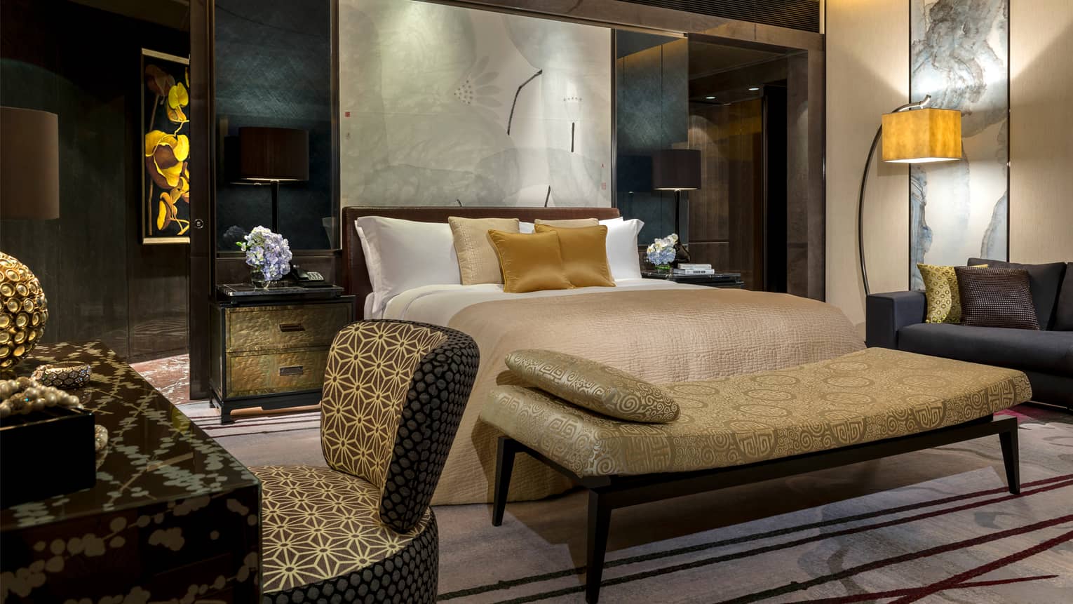Hotel room at night with dark brown and cream decor, gold accent pillows and bench, modern art