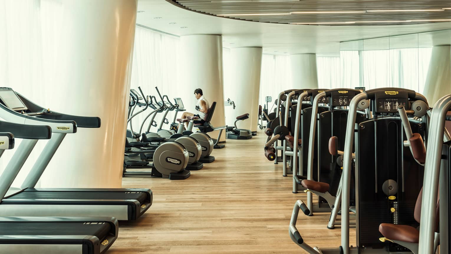 Cardio machines in bright fitness room with modern white pillars, man on bicycle machine