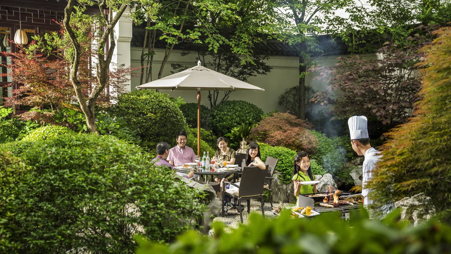 View over hedges, trees to family dining on patio, chef at barbecue