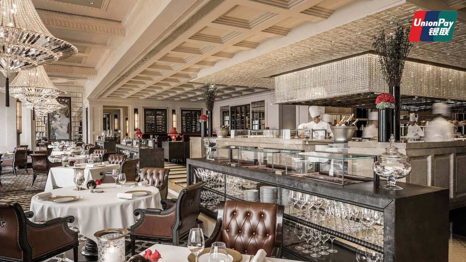 An elegant French restaurant interior with polished wooden floors, white tablecloth-covered tables, and plush leather chairs. The room features chandeliers. 