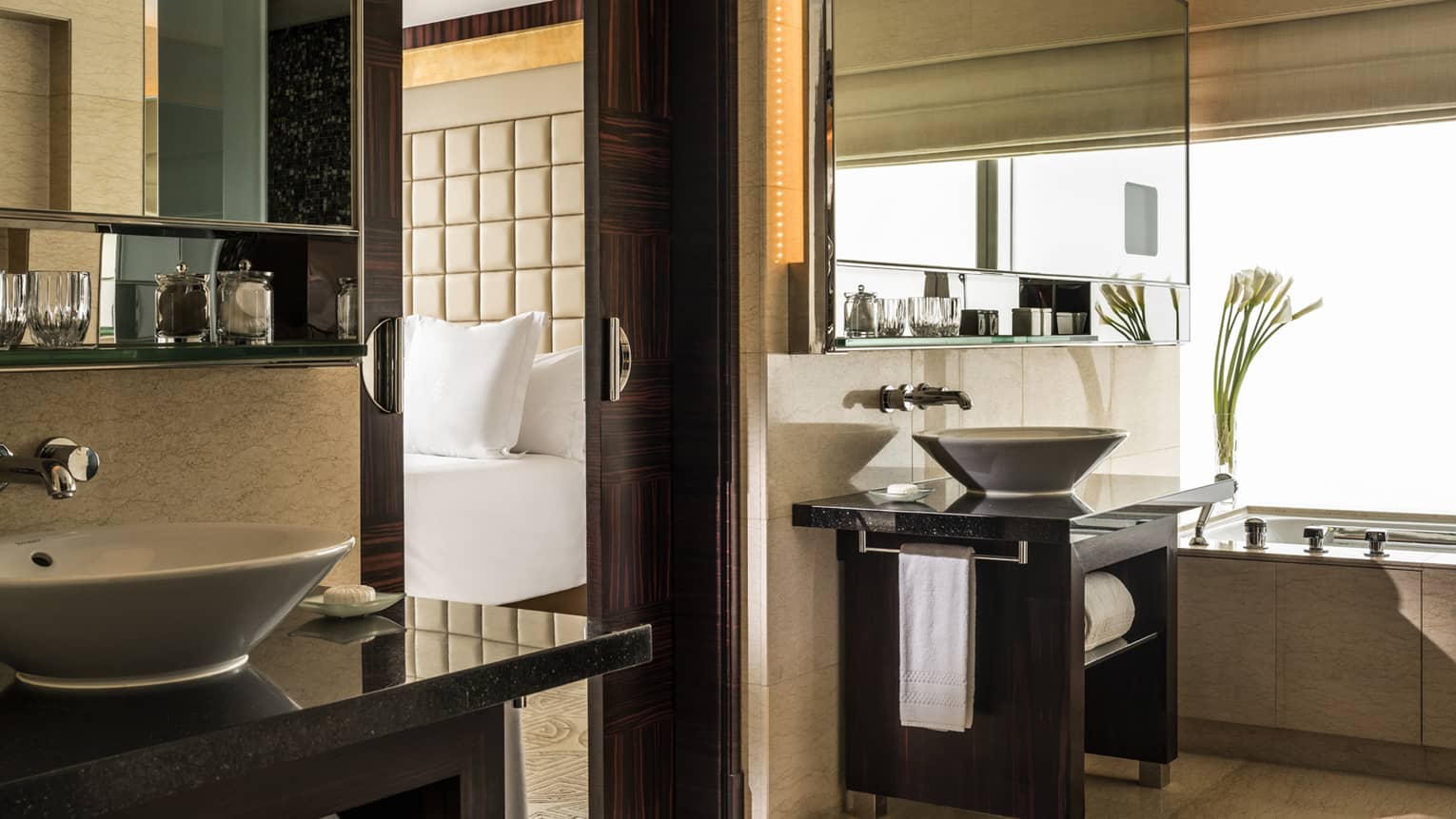 Elegant hotel bathroom with vessel sink, dark wood accents, large mirror, and glimpse of bedroom with tufted headboard.