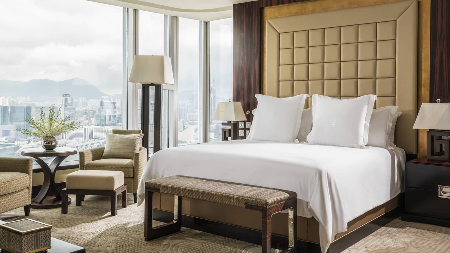 Luxury hotel bedroom with king-size bed, beige tufted headboard, city view windows, armchair with ottoman and decorative flower arrangement.