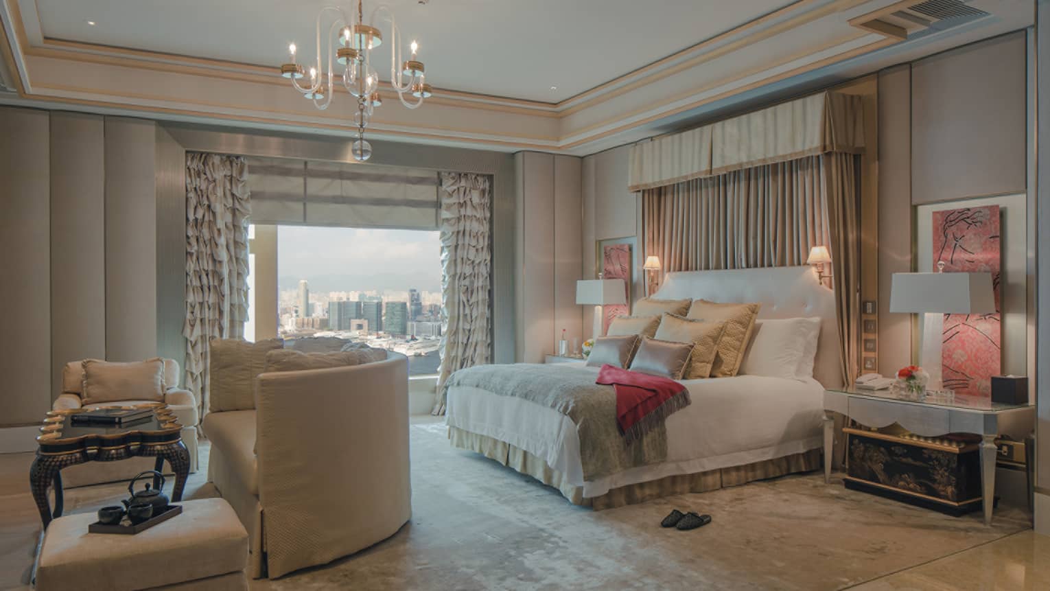 Luxurious bedroom with a large bed, plush pillows, and elegant drapery. A seating area is near the window, which offers a city view.