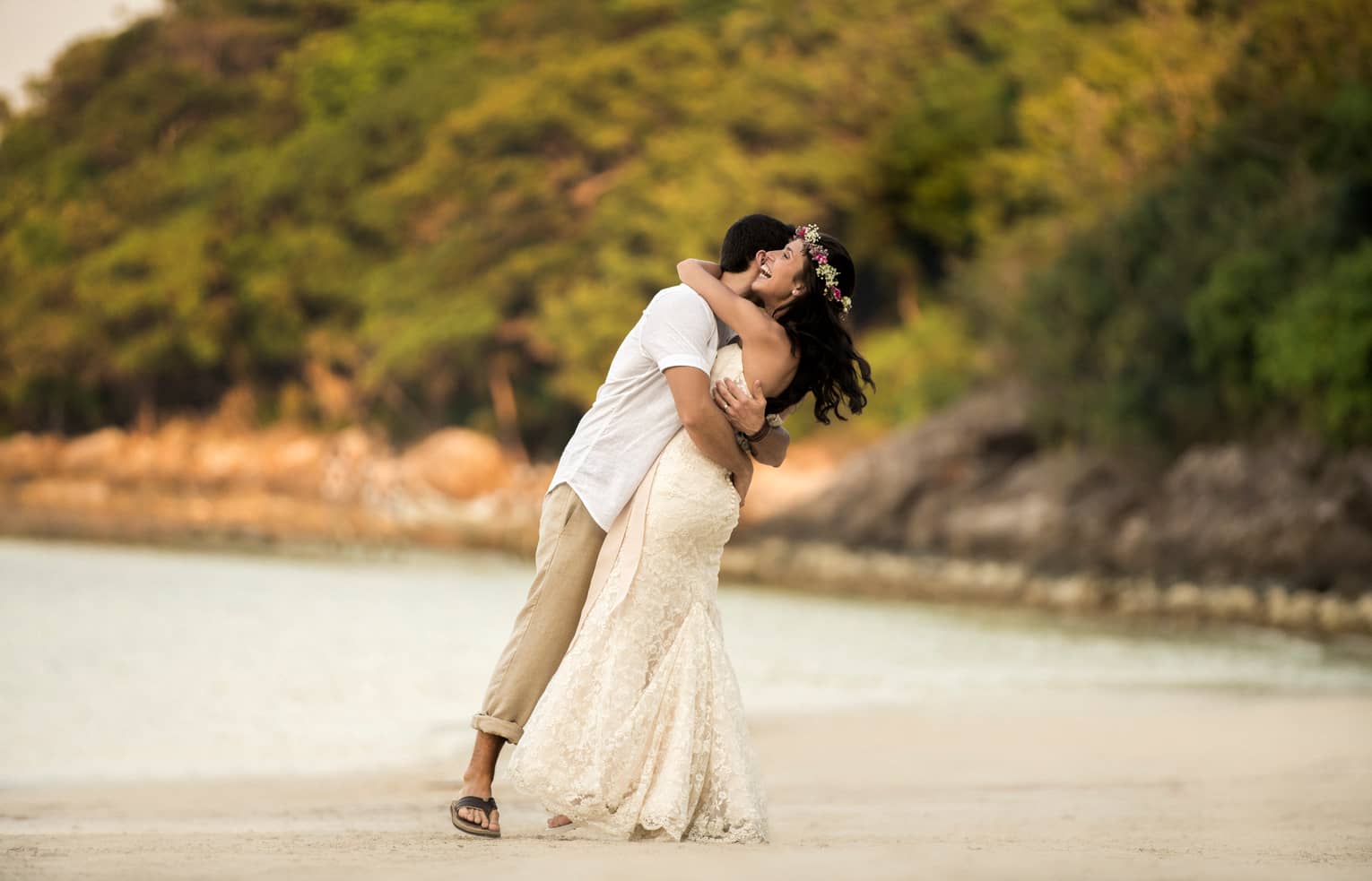 Man in white shirt and khaki pants, woman in long white wedding gown and flower crown embrace on the beach