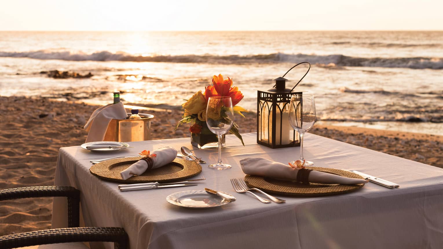 Close-up of formal place setting on table with white cloth, Champagne in bucket on beach at sunset