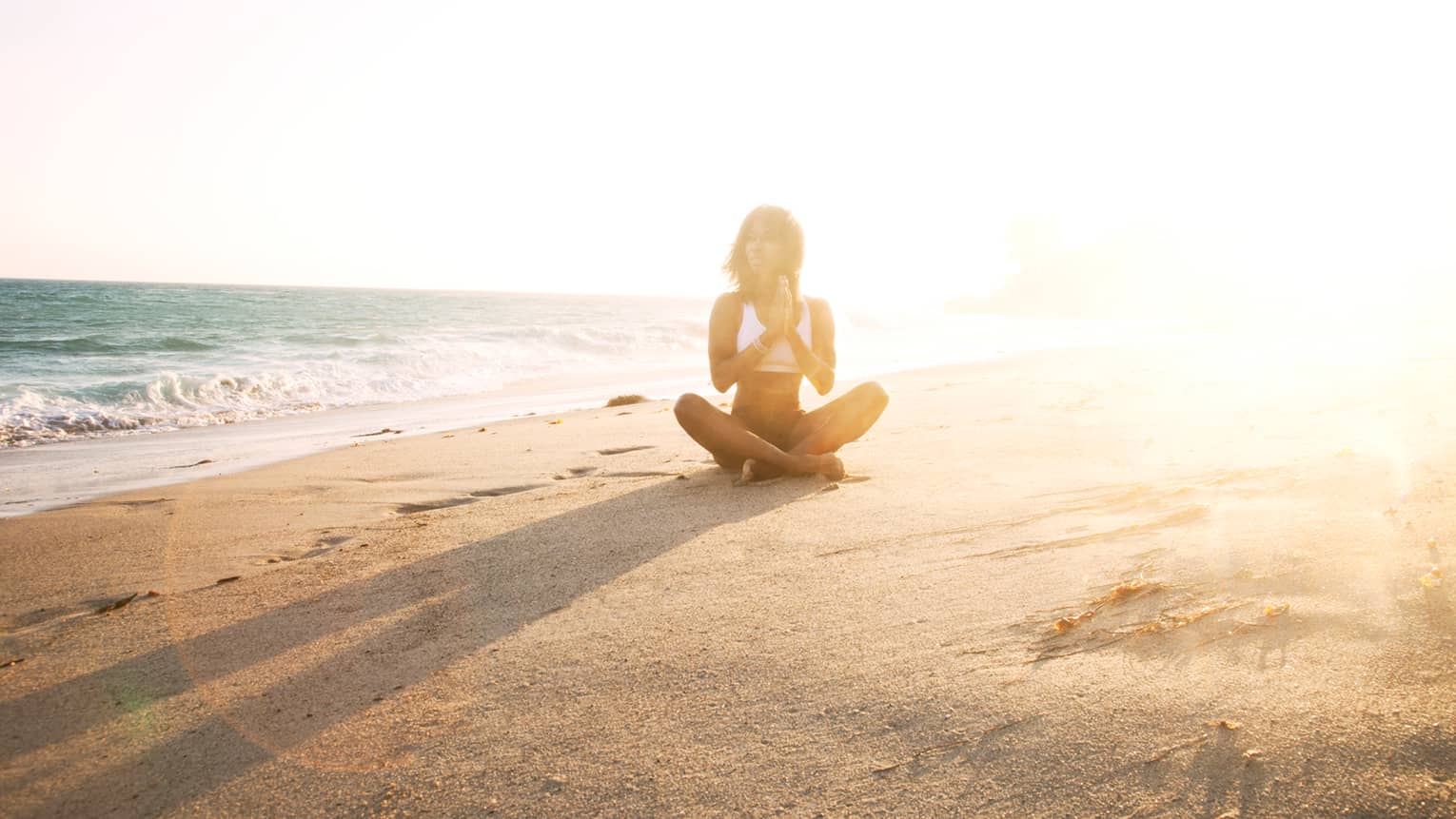 Woman in swimsuit sits cross-legged on sand beach at sunset