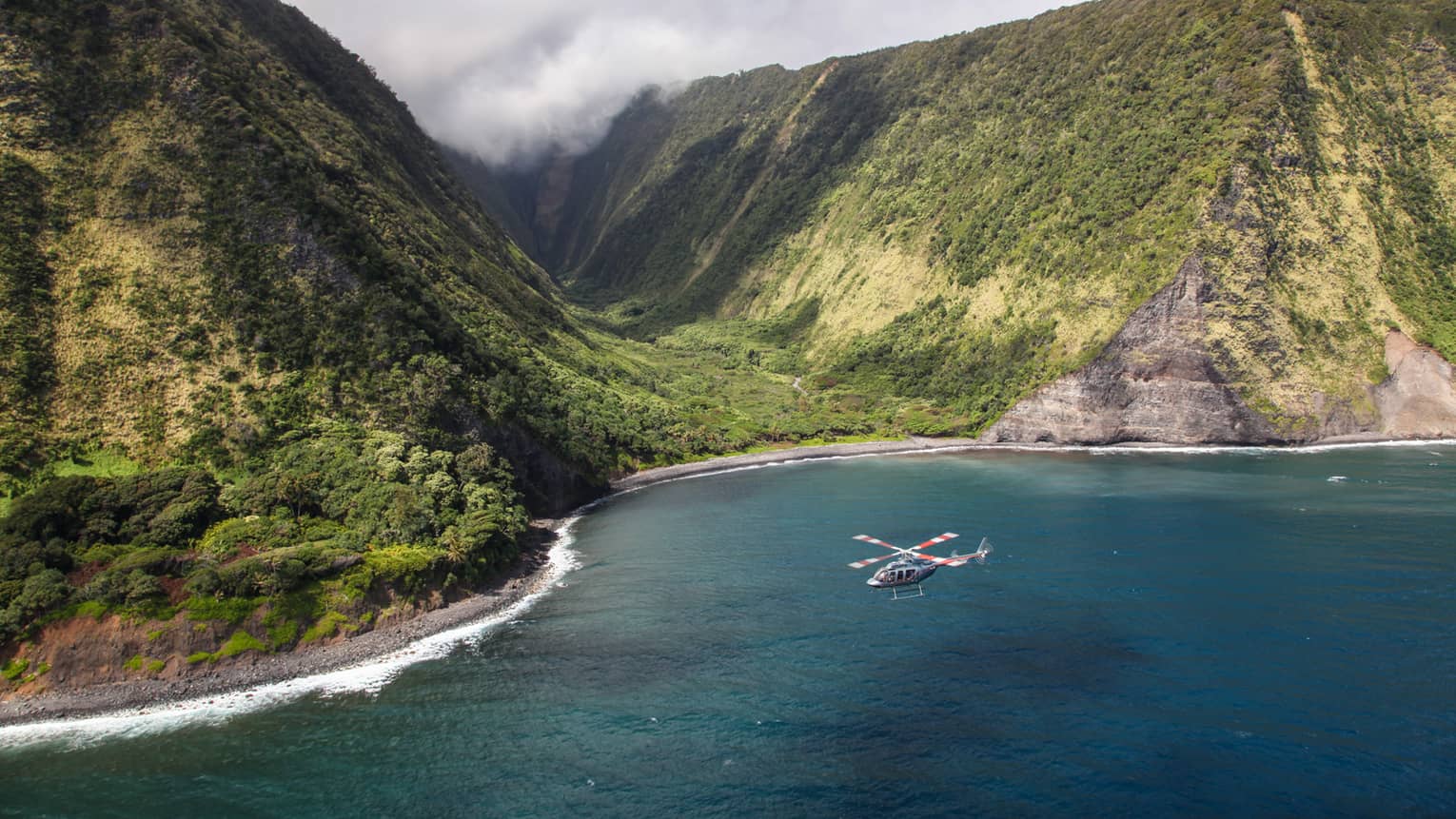 Helicopter flies over ocean by sweeping green volcanic mountains