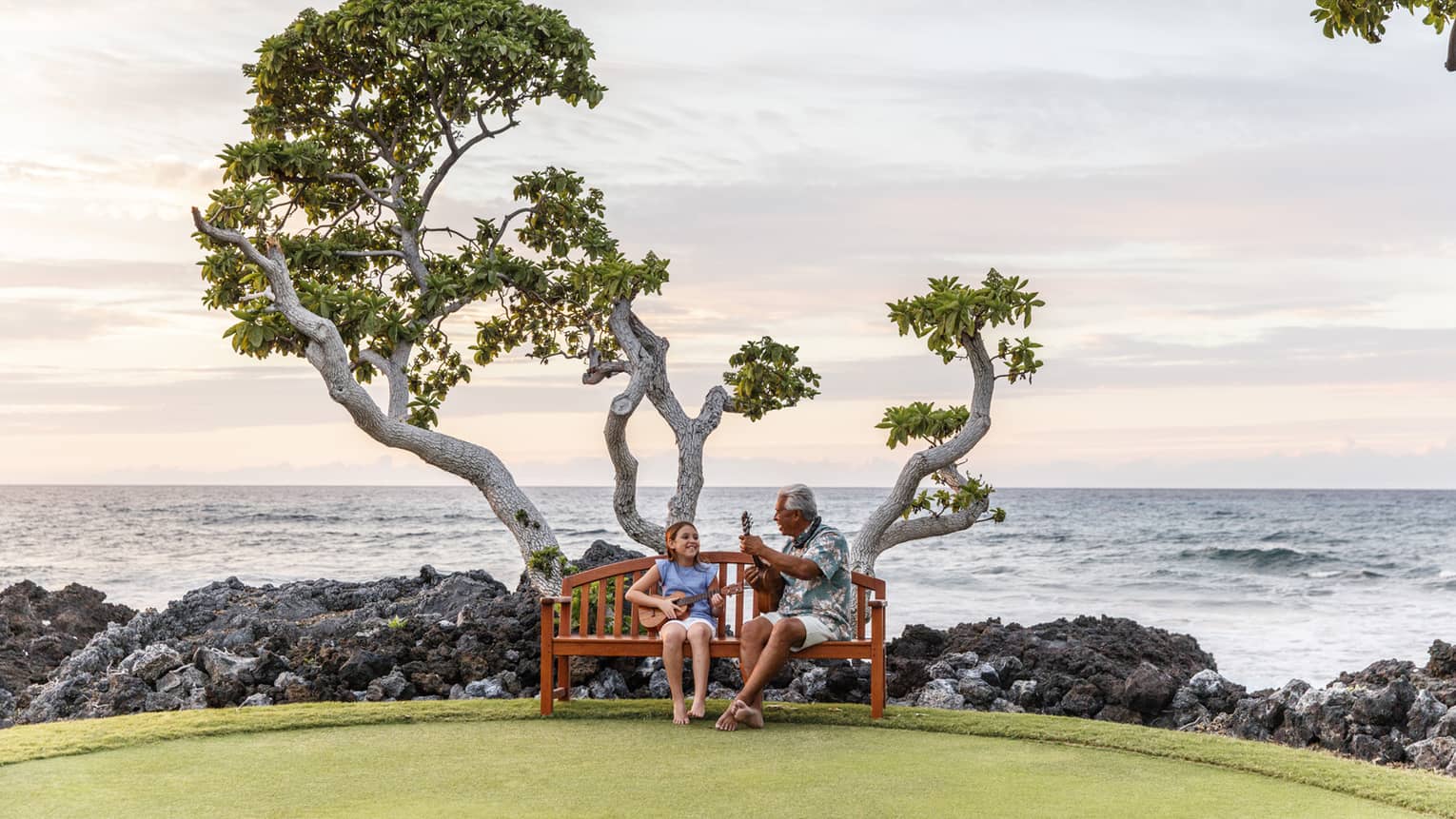 Hawaiian elder and young girl play ukuleles on bench under tree by black rocks, ocean
