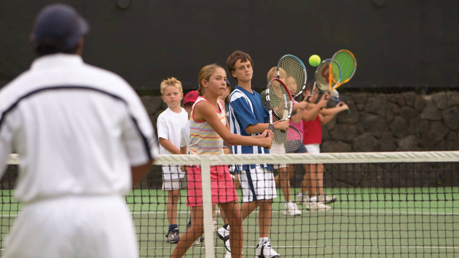 Tennis coach in front of group of kids playing tennis behind net