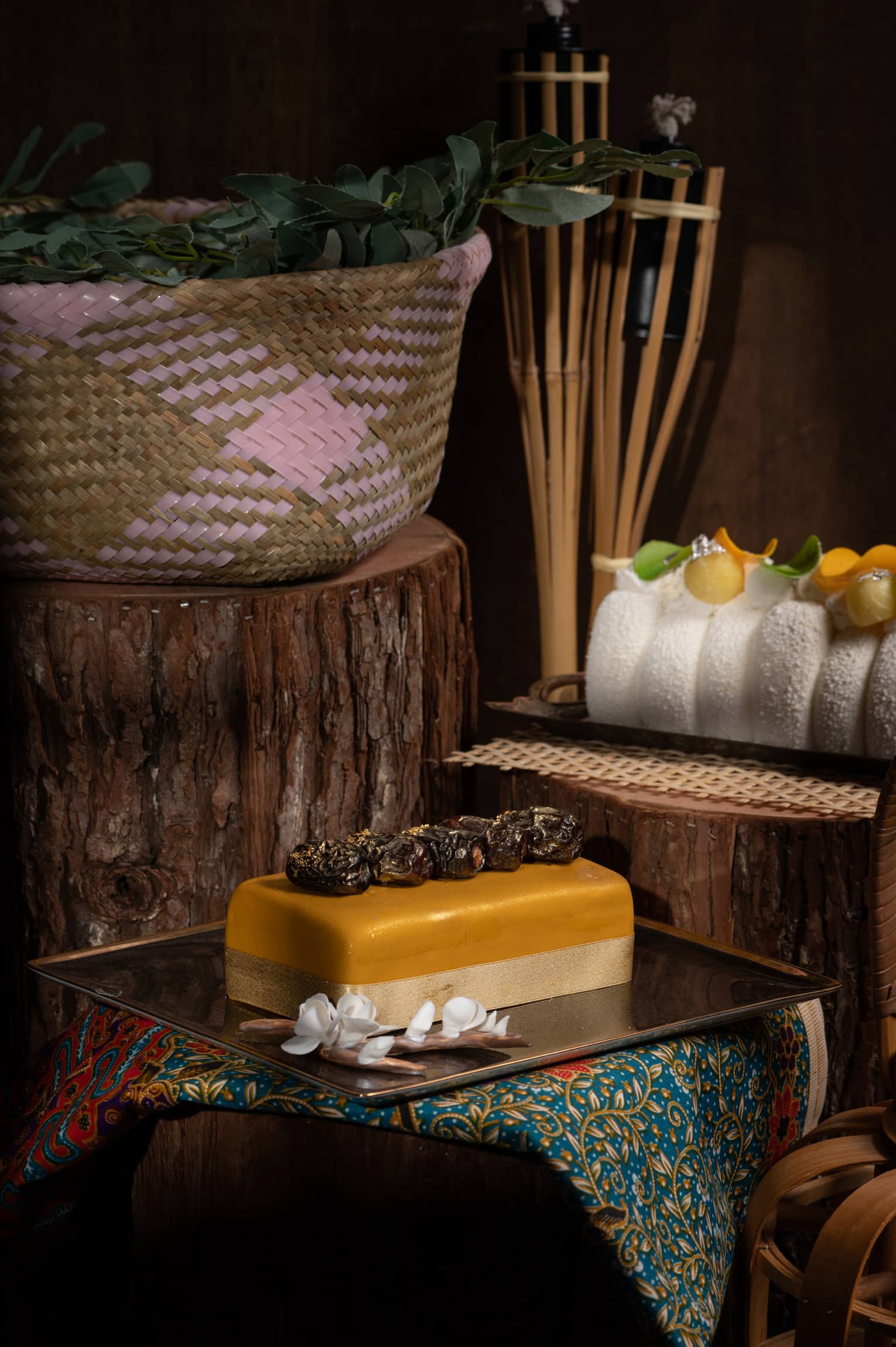 Four Seasons Raya cakes on pounded copper platters with white flower garnishes, woven pink basket of lime leaves on tree stump, tiki torches