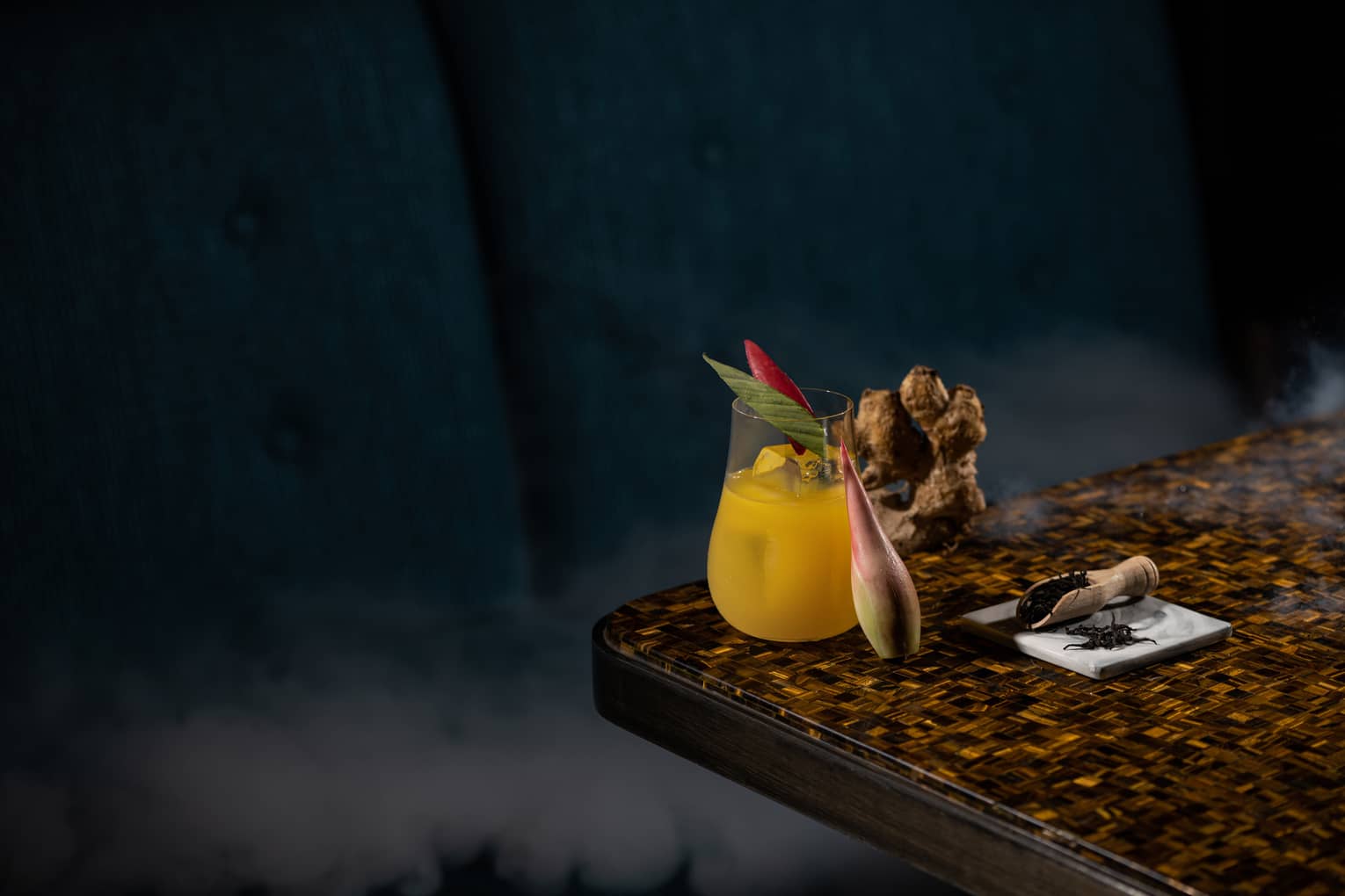 Kunyit Royale signature cocktail in stemless glass on edge of countertop with turmeric root and leaf garnish